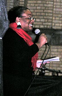 50ish woman in black, with a bright red scarf, large black-and-white earrings and glasses, smiling and holding a microphone in one hand and a sheet of paper in the other