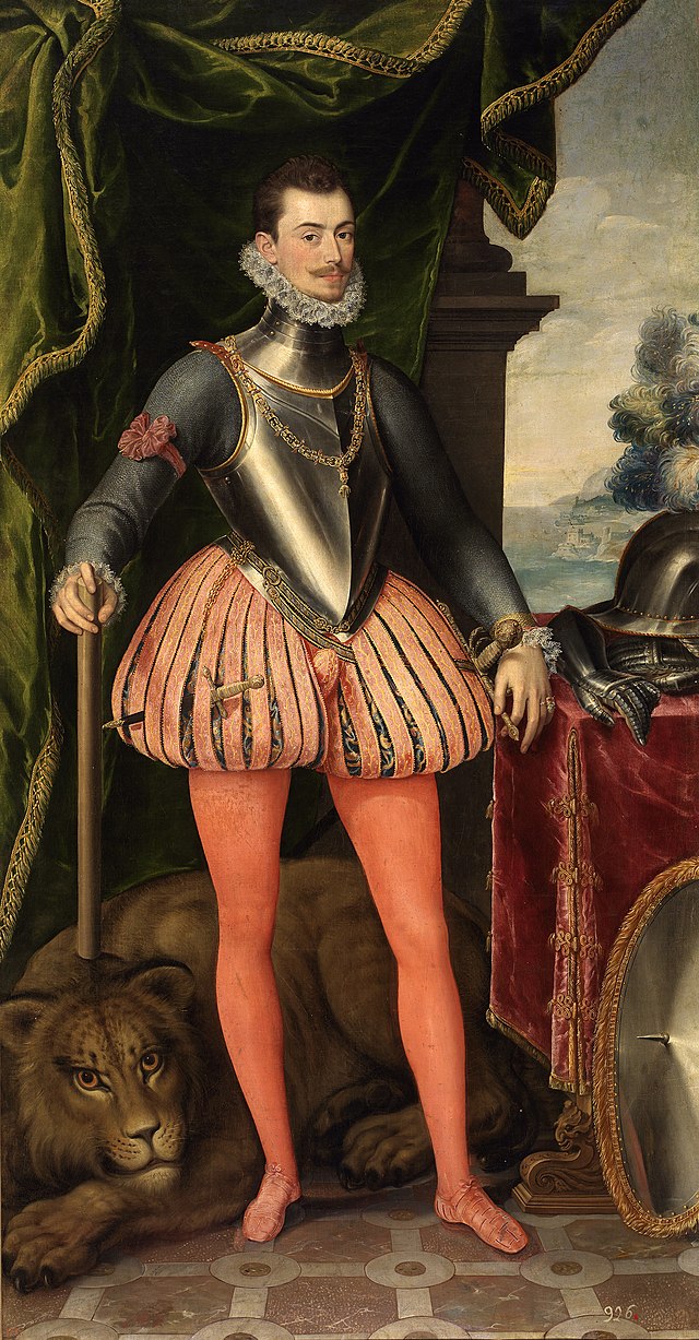  A tall brown-haired  man in armour and a royal attire posing for a painting.  He is standing under a luxurious green drapery and wearing a red brassard on his right arm.  An attentive lion is lying behind him at his feet.  His helmet and gloves are lying on a table beside him while his shield is resting on the floor in front of the table.