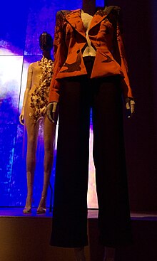In the foreground, an orange jacket with black pants. In the background, a sheer feathered dress. Both are on mannequins.