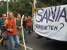 A group (about fifteen people are visible) composed predominantly of white males on a street. A large white handwritten banner is visible which reads "SALVIA VERBIETEN?" in large black letters.