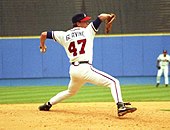 A left-handed baseball pitcher wearing a black baseball cap, white uniform, and black shoes; the back of his uniform has the lettering "GLAVINE" and the number 47, in a throwing stance.