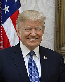 Official White House presidential portrait. Head shot of Trump smiling in front of the U.S. flag, wearing a dark blue suit jacket with American flag lapel pin, white shirt, and light blue necktie.