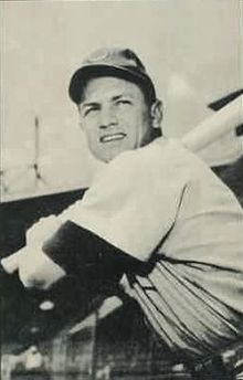 A in a light baseball jersey and dark cap posed prior to swinging his baseball bat