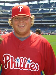 A smiling young man with feathered blonde hair wearing a red baseball jersey and cap