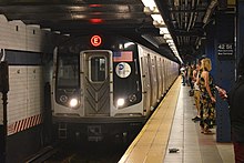 The front end of a subway train, with a red E on a LED display on the top. To the right of the train is a platform with a group of people waiting for their train.