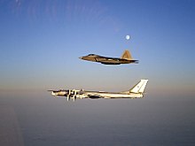 Aerial port view of two aircraft in flight, one on top of the other. The bottom aircraft is a four-engined propeller-driven aircraft, which is escorted by a jet fighter. The Moon is visible as a tiny spot in the sky.