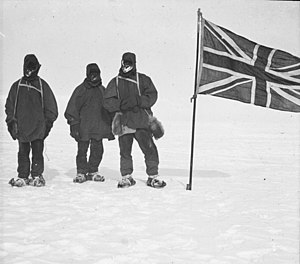 Three men in heavy clothing stand in line on an icy surface, next to a flagstaff from which flies the flag of the United Kingdom of Great Britain and Ireland