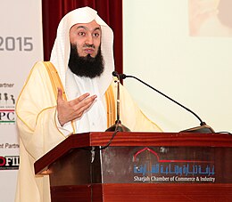 Homme barbu en tenue traditionnelle musulmane blanche et dorée devant un podium avec un micro et le bras droit levé.