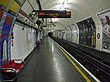 The interior of a building with rounded ceiling and walls, red benches along the left wall, advertisements along the right wall, and a yellow line on the floor