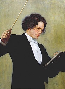 A middle-aged man with long dark hair, wearing a tuxedo and standing behind a music stand, waving a conductor's baton.