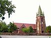 A church with a red roof, and red body with yellow bands, the tower is to the right with a spire, four pinnacles and a clock face; in front is a wall and a surfaced car park