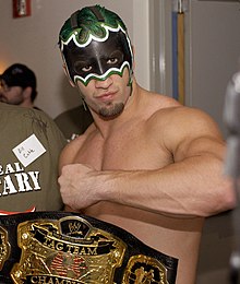 A topless Caucasian man, with green hair and wearing a black and white mask that covers the top half of his face, poses with his arm rasied across his chest and his hand in a fist. A wrestling championship with a black strap is visible in the foreground of the image, and is inscribed with "WWE Tag Team Championship".