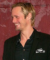 A man with blond hair and dressed in a black shirt and blazer smiles to his right