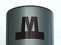 A photograph of a rounded grey sign with small, circular, evenly spaced, linearly organized perforations and a large "M" written in dark red