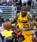 Black man dribbling a basketball with a gold and maroon basketball jersey that says Cavaliers in maroon cursive lettering across the chest. He is wearing a headband with an NBA logo.