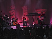 A woman singing on a microphone, standing up, with a band with instruments in her back and a crowd of people in front. There are 3D letters in the background, spelling "Lily".