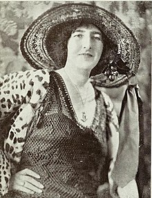 A middle-aged white woman, smiling, wearing a wide-brimmed hat and a leopard-print jacket. She has her hands on her waist.