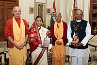 A color photo of the President flanked by Hare Krishna followers in robes, holding a copy of the Bhagavad Gita in Russian.