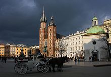 Zdjęcie przedstawiające fragment krakowskiego Rynku. Przed kościołem świętego Wojciecha z białego kamienia wapiennego, z kopułą pokrytą spatynowaną miedzią stoi dorożka. Nie jest to typowa krakowska dorożka, a raczej biała dworska karoca z zaprzęgniętymi dwoma końmi. Za kościołem wzdłuż Rynku ciągnie się szereg kamienic. W rogu wznosi się ceglany Kościół Mariacki z dwoma wieżami. Wyższa wieża zwana Hejnalicą ( z niej rozbrzmiewa hejnał) została zbudowana na planie kwadratu, od dziewiątej kondygnacji przechodzi w ośmiobok jest zwieńczona gotyckim hełmem składającym się z ośmiobocznej, zaostrzonej iglicy, otoczonej wieńcem ośmiu niższych wieżyczek. Niższa wieża jest kwadratowa, zwieńczona późnorenesansowym hełmem składającym się z osadzonej na ośmiobocznym bębnie eliptycznej kopuły zwieńczonej ażurową latarnią. W narożnikach ustawione są cztery mniejsze kopułki na niskich, sześciobocznych podstawach. Pośrodku tej części Rynku stoi pomnik Adama Mickiewicza, a wzdłuż linii A-B widać rozstawione żółte parasole straganów krakowskich kwiaciarek. Niebo nad Rynkiem jest ciemnogranatowe.