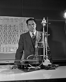 Photographie en noir et blanc de Glenn Theodore Seaborg de face derrière un montage de chimie avec un tableau périodique en arrière-plan.