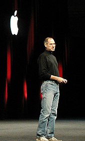 Full-length portrait of man about fifty wearing jeans and a black turtleneck shirt, standing in front of a dark curtain with a white Apple logo
