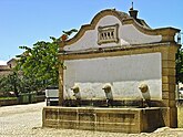 A public fountain in a town square, with three brown metal nozzles stuck evenly into a white wall.