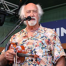 White-haired Michael Doucet holding a fiddle and bow in his right hand