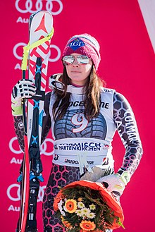 Femme en combinaison de ski à dominante grise, portant des lunettes de ski à effet miroir, un ski appuyé contre son épaule droite