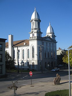 A formal building with arched windows and two domed towers is on a street corner near other less formal buildings. A car is parked in front of the formal building. Diagonally opposite are a stop sign and a small stone sculpture or monument.