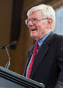 A Caucasian man with thin white hair. He is visibly older, with a somewhat wrinkled face. He wears round glasses, a blue button up shirt, a red necktie, and a black suit jacket. He stands at a podium with two microphones in front of him. He is in the middle of speaking.