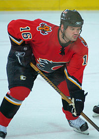 Hockey player in red uniform. He crouches, his feet shoulder width apart, holding