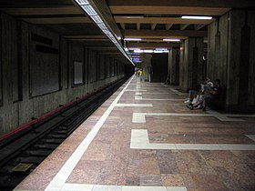 Une partie du quai central, le sol est en marbre rouge avec des motifs géométriques blancs, et une voie du métro