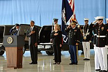 Обама и Клинтон на мрачном мероприятии, чествуя жертв нападения в Бенгази на церемонии передачи останков, состоявшейся на базе ВВС Эндрюс 14 сентября 2012 года. Солдаты стоят позади Обамы и Клинтона, и все стоят на большом деревянном полу с их левые руки по бокам и их правые руки на верхней части груди.