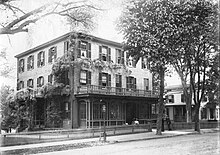 A three-story building on which a flowering vine is climbing on the left behind a low fence with a tree on the right