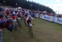 Peloton de plusieurs dizaines de coureurs à VTT avec en arrière-plan un public derrière une barrière.