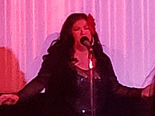 Photographie d’une femme qui chante devant un micro, habillée en noir.