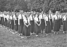 Photographie en noir et blanc d'un groupe de jeunes filles debout en uniforme et chemise blanche. Disposées géométriquement, elles se tiennent au repos.