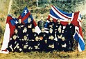 Photograph of the Native football team in their uniform of black shorts and jerseys sat in front of the flags of Great Britain and the United Tribes of New Zealand