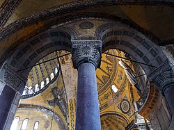 A cropped photograph taken from the ground level of the interior of a building built in the classical style. Arches and pillars painted blue, and ornate walls painted yellow can be seen, all of them are proportional