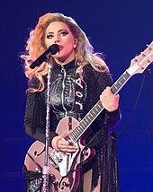 Lady Gaga standing behind a microphone stand with a pink guitar in her hands, wearing black leather fringe.