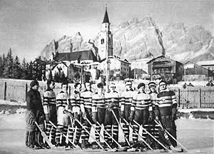 Eleven men, dressed in sweaters and skates while holding ice hockey sticks, stand on ice outdoors, with a large building in the background.