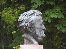 The grey scuplture of a head of a man in his sixties on a plinth with trees in the background. The front of his face is clean shaven but sideburns run under his chin.