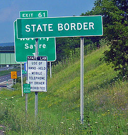 Бело-зеленый знак с надписью «Государственная граница». Позади него вдали виден стандартный знак выезда с автострады США.