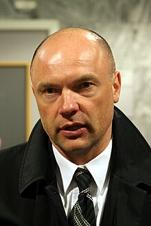 A photograph of a man at a press interview. He is wearing a black coat and a white shirt. The man is in the middle of a discussion with a person not seen in the image.