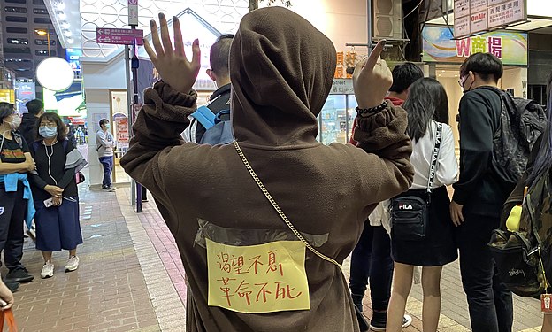 香港市民阿澧11月30日晚在旺角街站寫心意卡給12港人，她在背上貼上標語，高舉反送中運動」五大訴求、缺一不可」的手勢。