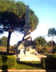 Photographie d'un monument avec une forme semblable à celle d'un obélisque et avec un aigle en bronze sculpté haut-relief sur son socle. Le monument est situé dans un jardin avec un pin derrière.
