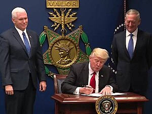 Donald Trump signing the order in front of a large replica of a USAF Medal of Honor, with Mike Pence and James Mattis at his side