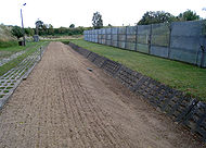 View along a ploughed bare earth strip, the right-hand side of which descends into a ditch faced with concrete. A metal fence is situated further to the right.