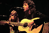 Мужчина с длинными черными волосами играет на гитаре с микрофоном перед ртом. Вдалеке женщина смотрит прямо в камеру.