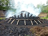 An aerial view of a bearded man in a coat, with a handmade rake, standing on a low, ~10m wide, rounded mound of black earth. The mound is emitting many slender streamers of smoke from all of its surface except the bottommost meter. The steamers merge and stream diagonally away from the viewer on a gentle but chill morning breeze, making an opaques white cloud. The visible background is dark coniferous forest; in the foregroud, a glimps of another pile, this one tightly-packed wood not yet covered with soil.